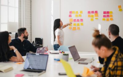 a leader at whiteboard with sticky notes talking to mixed group of people in foreground sitting at a conference table